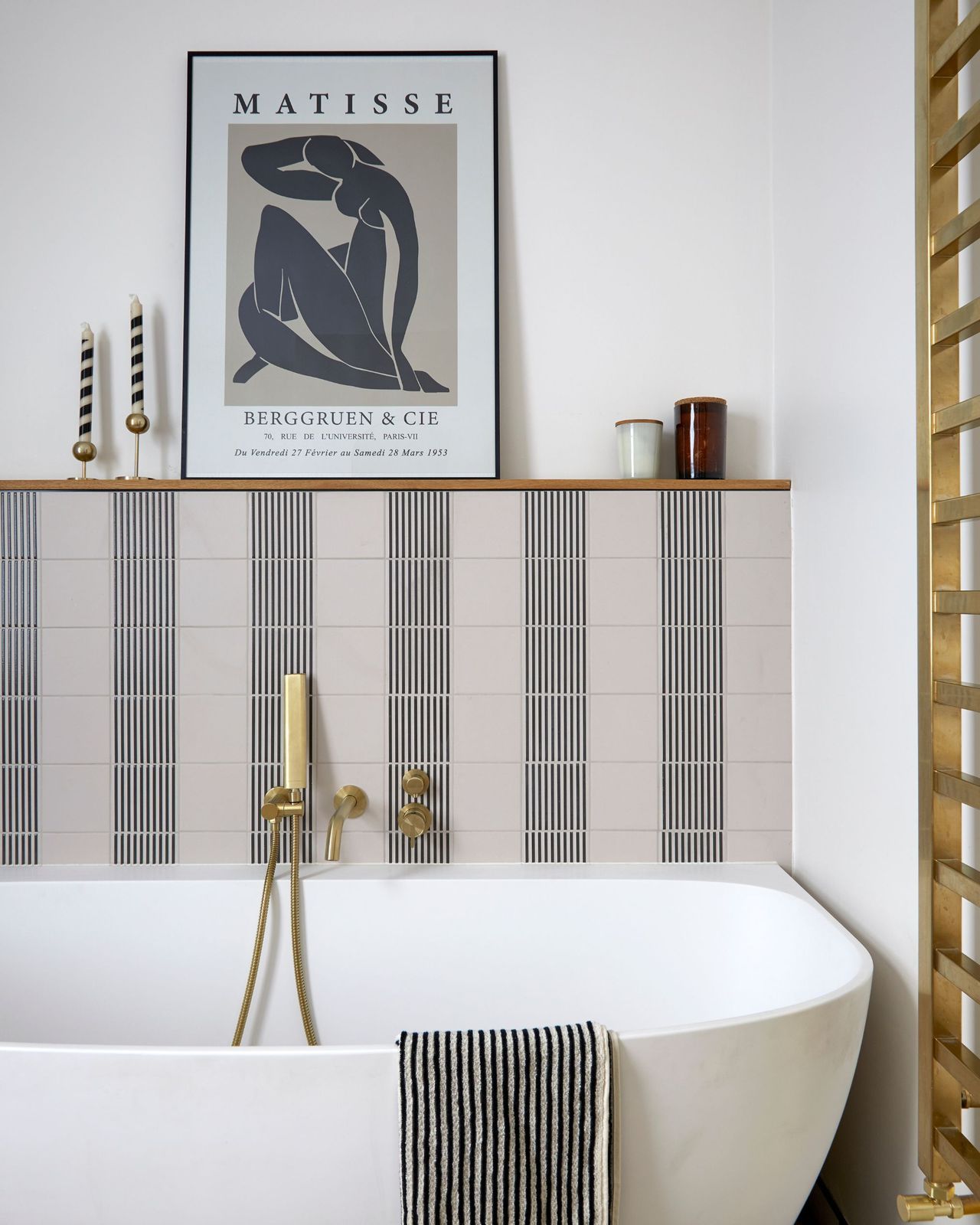 A modern bathroom with a white bathtub and gold radiator with a shelf above tiles with a framed print and candles