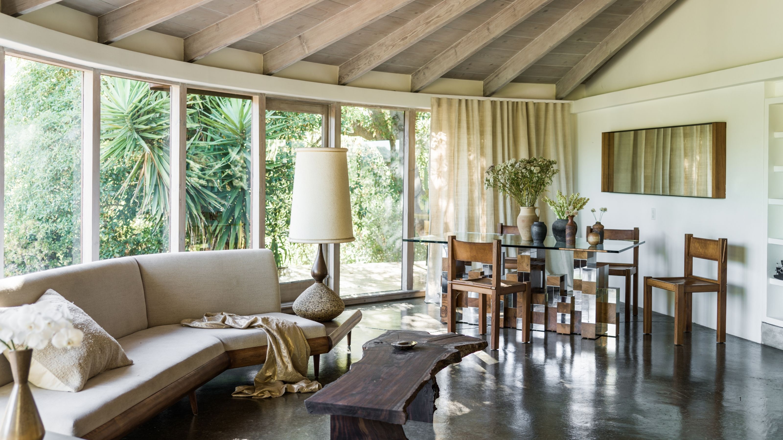A modern room with a curved window that has linen, beige curtains. Wooden ceilings, and a beige sofa with a rustic coffee table in front of it. A dining table with leather and wood dining chairs