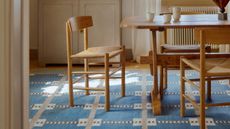 Image of a modern rustic dining room with a wooden table and chair set, styled on top of a blue, geometric area rug. There is a window with white curtains on the back wall. Beside the window is a storage cupboard with dishes inside.