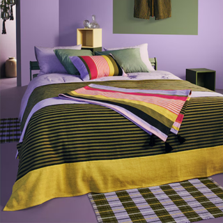 A bedroom with purple walls, green and purple striped bedding, and a green and purple rug