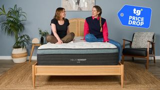 Mattress testers Ruth Jones and Nicola Appleton sit on the Helix Sunset Luxe mattress on a bed frame in the Tom's Guide Sleep Lab. A Tom's Guide Price Drop deals graphic in the top right corner