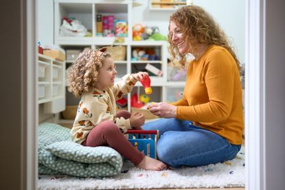 A nanny and a child are sitting on the floor in a room with a lot of toys