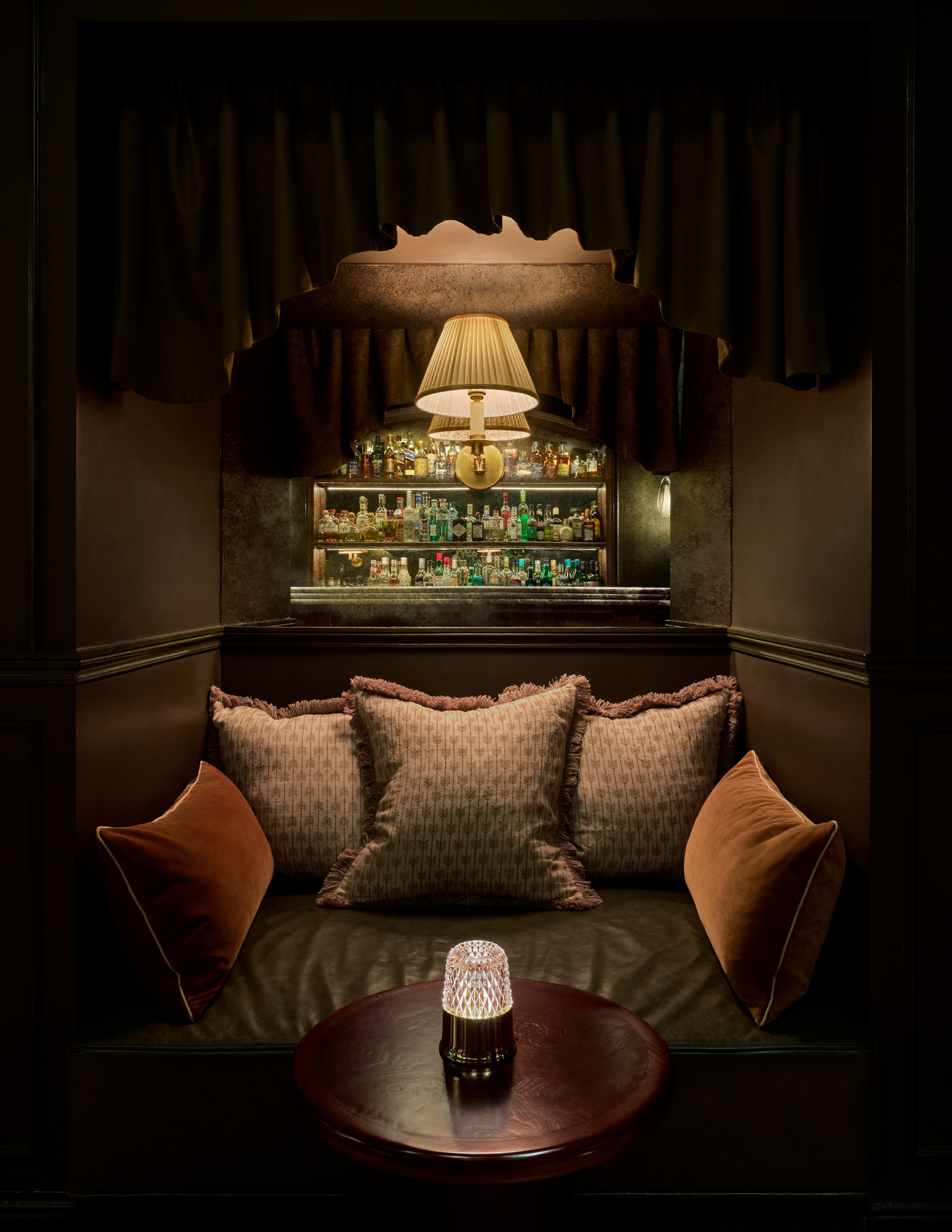 A love seat carved from dark green leather into a wooden column, with earthy-toned cushions, a golden lamp, a round table in wood, and a tabletop lamp, topped with a mirror reflecting a stocked bar.