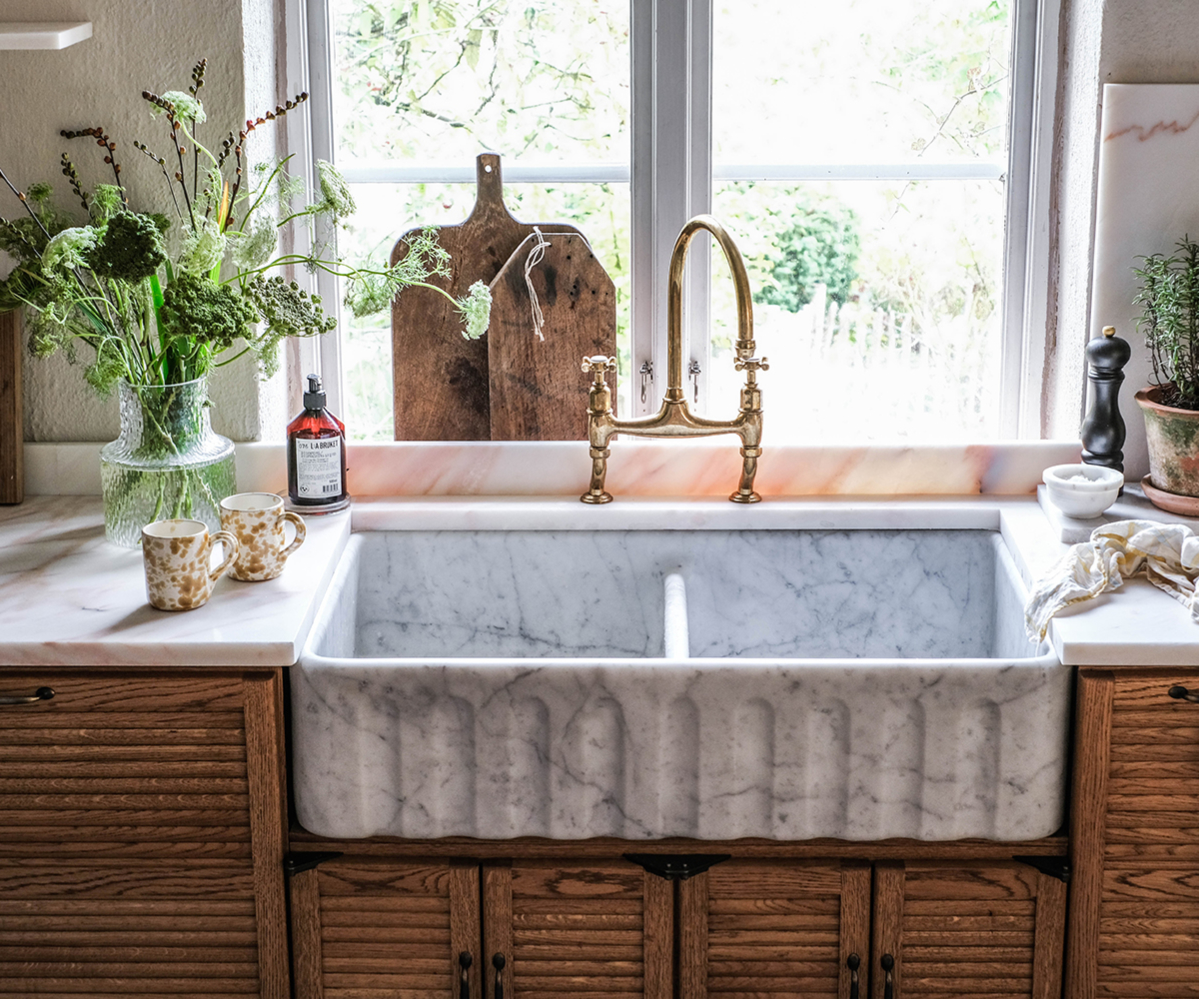 close up of white marble butler style kitchen sink set in wooden fronted cabinets in front of window