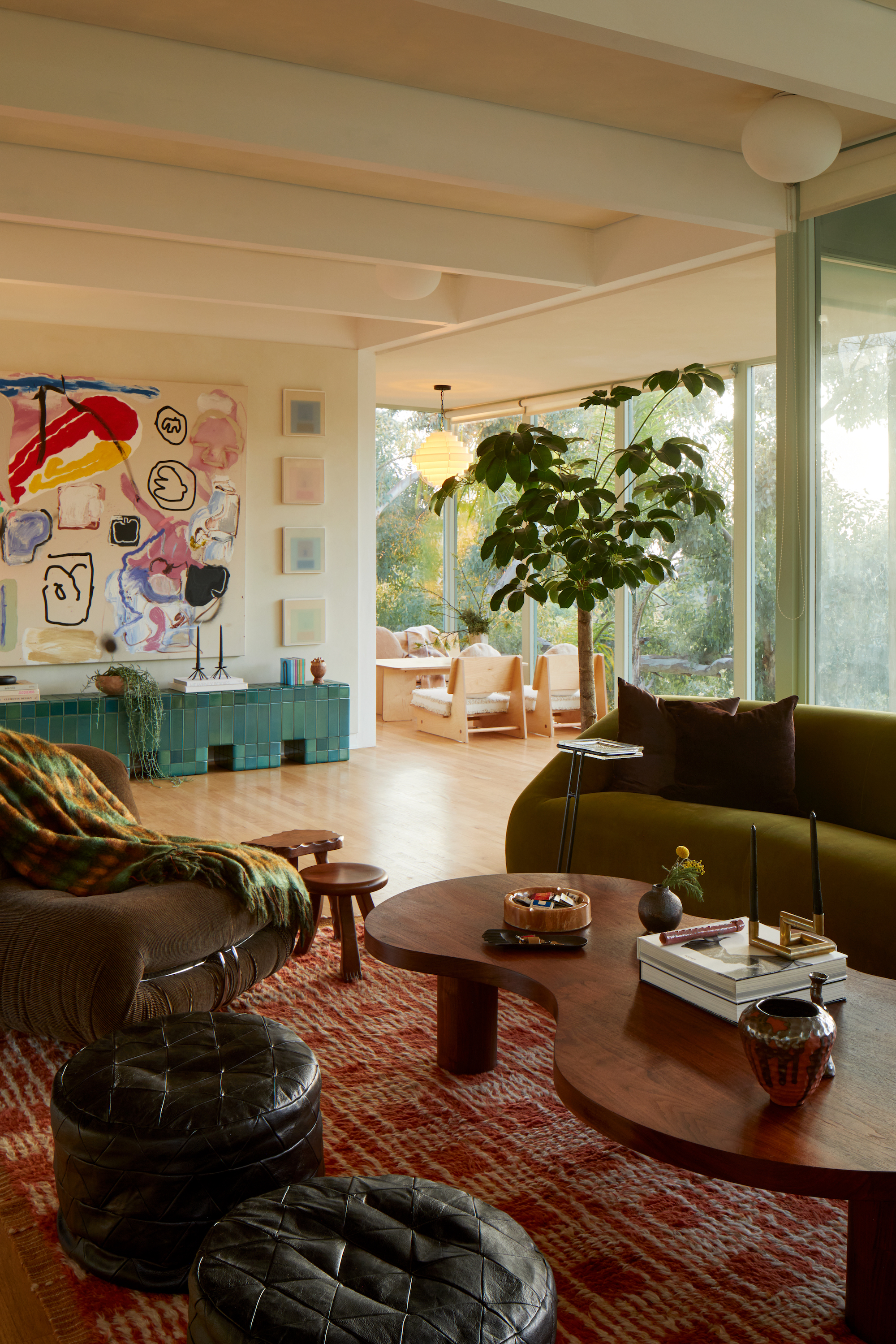 An open plan living area with floor to ceiling windows to the right, and an olive green sofa, dark wood coffee table and a bright artwork on the far wall.