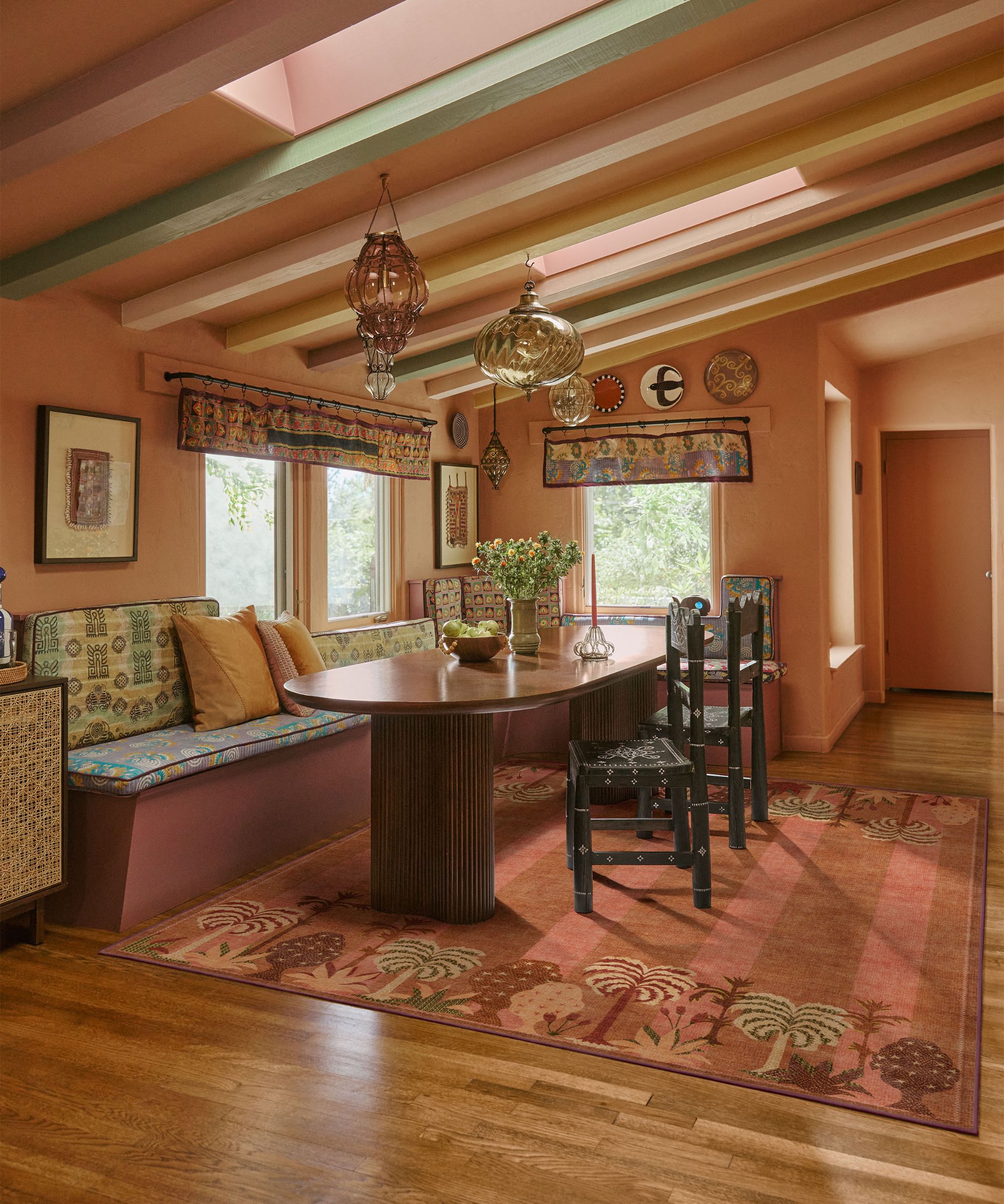 A peach painted dining nook with a built-in banquette with an upholstered seat, a curved wooden dining table, a colourful pink striped rug, patterned blinds, glass lantern lights, and a wooden floor.