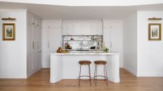 A white kitchen with gold hardware, a pair of brown stools, a marble backsplash and built-in shelving