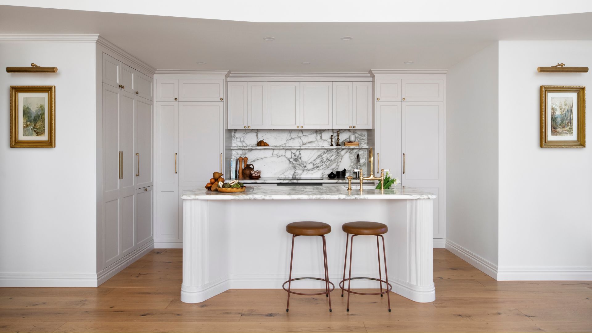 A white kitchen with gold hardware, a pair of brown stools, a marble backsplash and built-in shelving