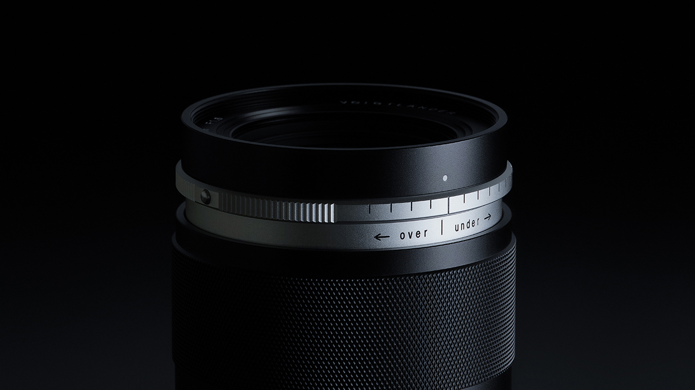 Close-up of a black camera lens with silver accents (the Voigtlander Portrait Heliar 75mm f/1.8) , featuring an adjustable AS control ring with &quot;over&quot; and &quot;under,&quot; options against a dark background