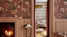 Living room with pink floral wallpaper, dusty pink panelling, a fireplace, a floral ottoman, looking into another living room with chevron wallpaper and a cream armchair.