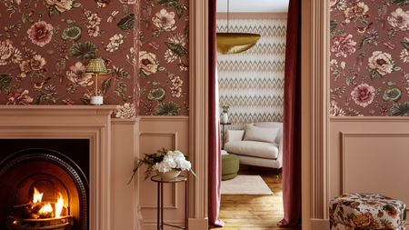 Living room with pink floral wallpaper, dusty pink panelling, a fireplace, a floral ottoman, looking into another living room with chevron wallpaper and a cream armchair.