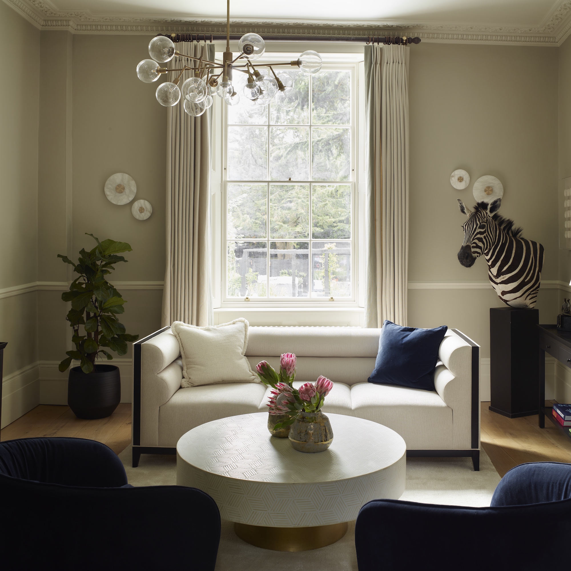 sitting room with cream walls, sash window, white sofa, round white coffee table and blue armchairs, with sputnik light and stuffed zebra