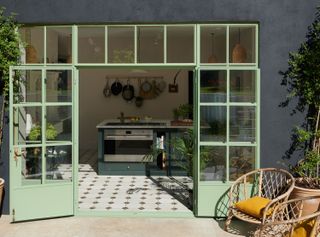 Pale green crittall windows with green kitchen, black and white tiled floor and rattan chairs.