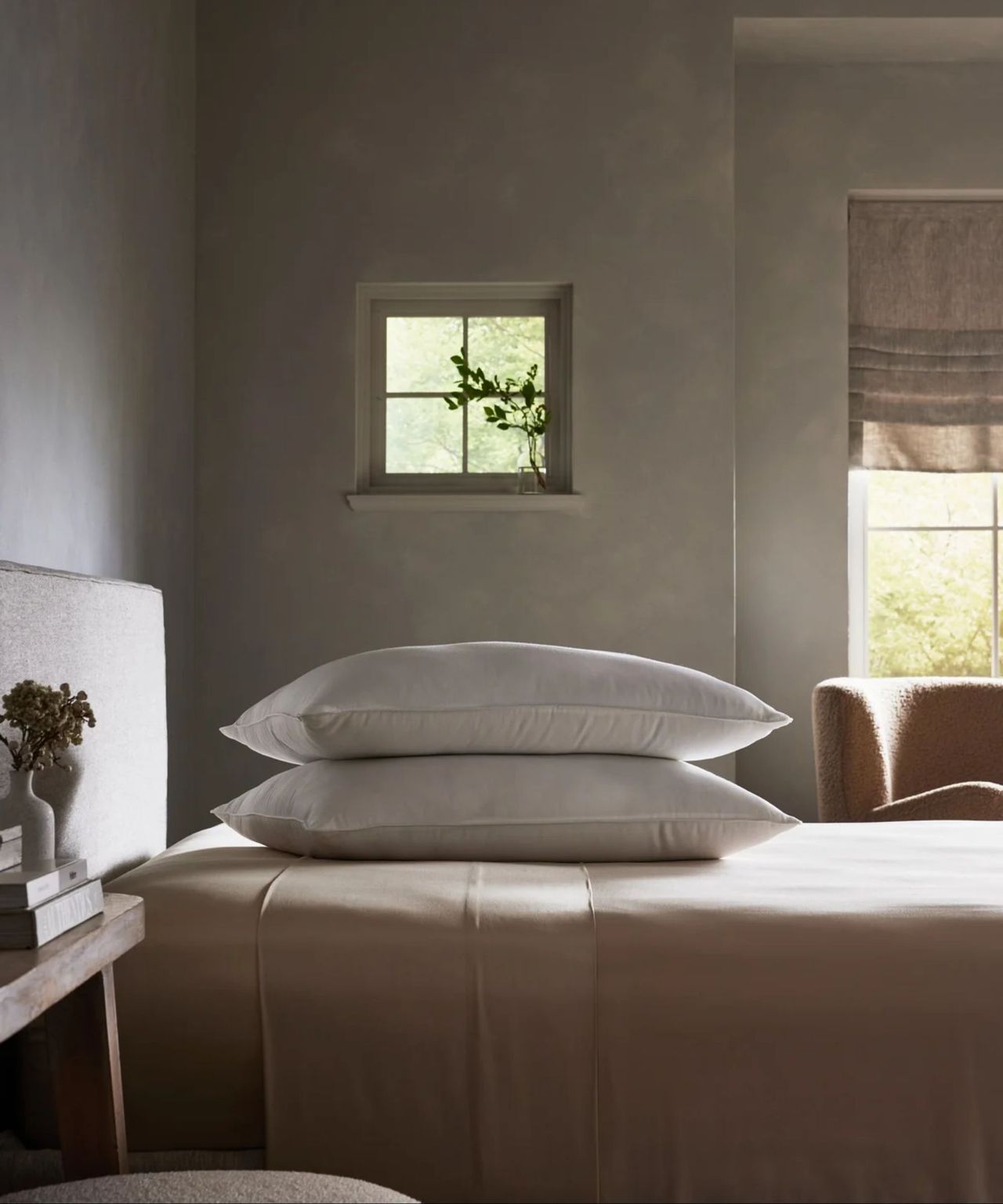 Cozy Earth Down Alternative Pillow pictured in a neutral bedroom with grey walls and a double bed with silk bedding. Behind are two windows, and in the foreground is a wooden nightstand with flowers.
