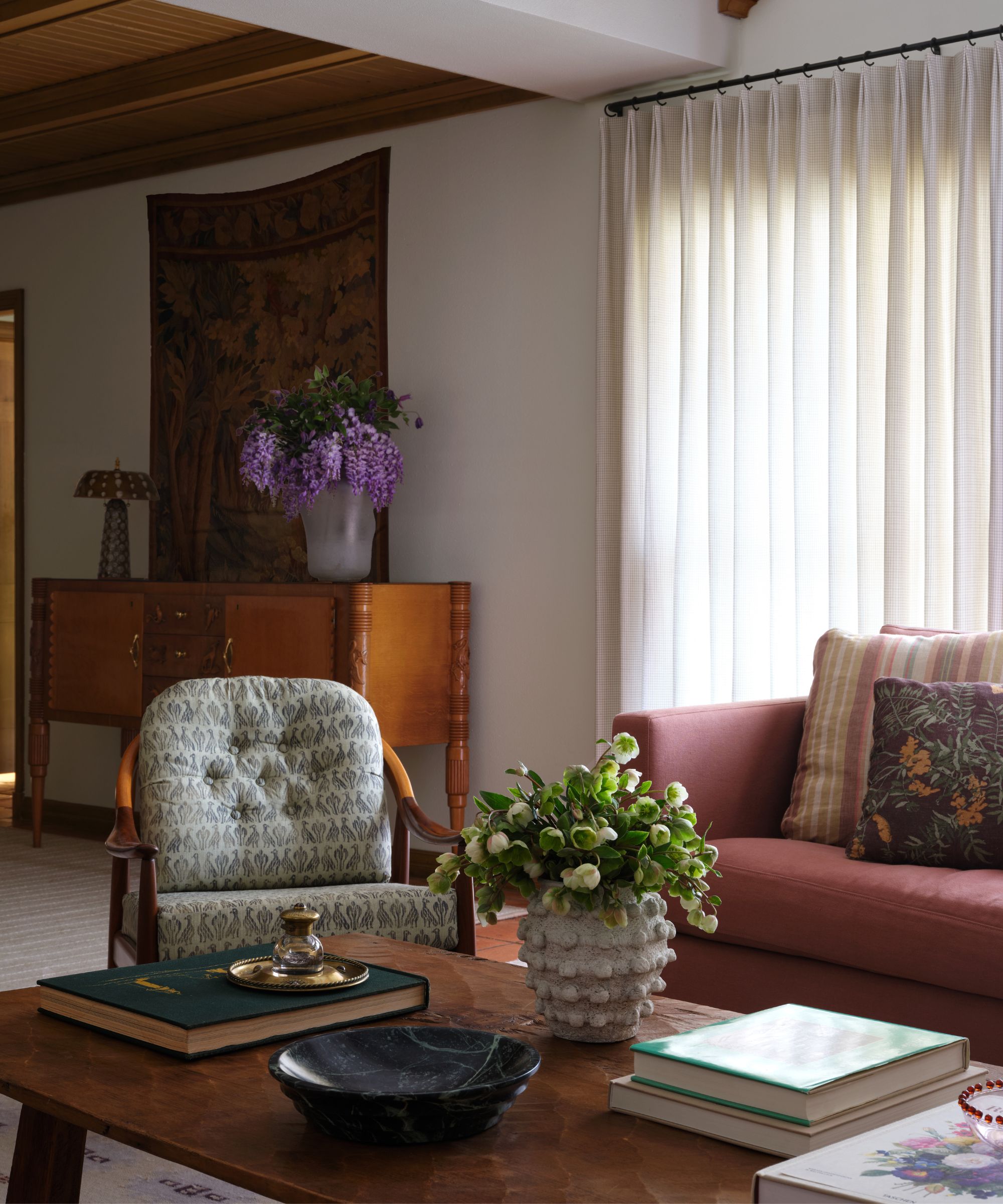 A living room detail showing a vintage wooden sideboard with purple flowers, a patterned armchair, and the edge of a pink sofa under soft window light