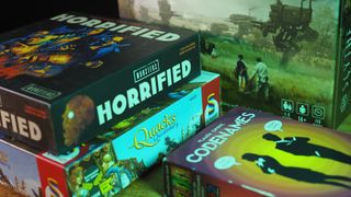 A stack of Horrified and Quacks of Quedlinburg board games beside Codenames and Scythe, all on a wooden table