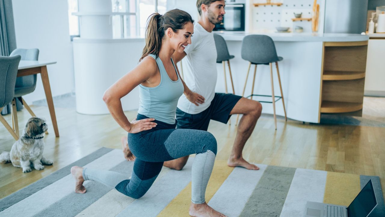 a woman and man in a home room setting both doing a split squat side on to the camera on a stripy rug and a small dog behind them.