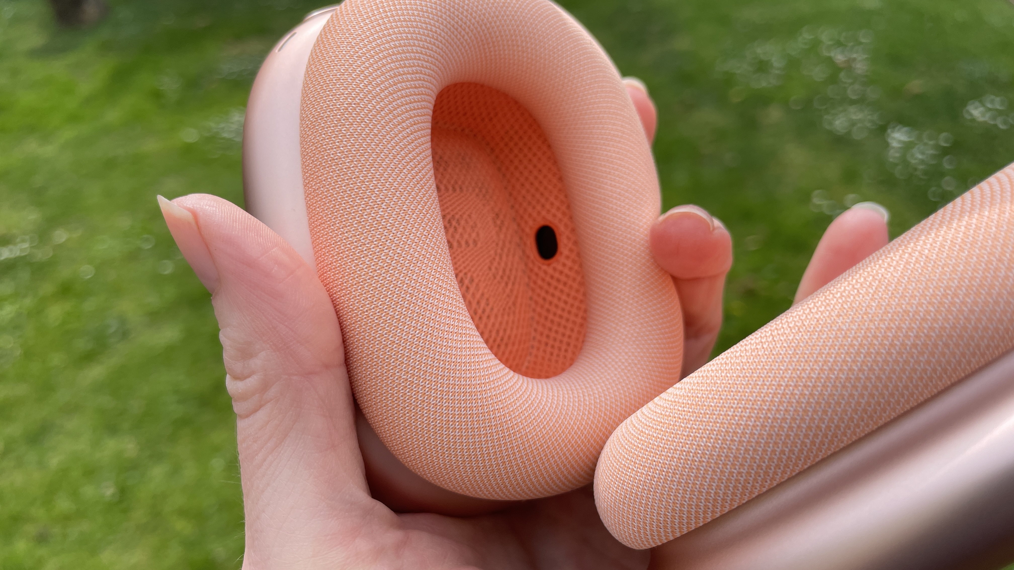 Apple AirPods Max 2 in orange with AirPods Max just visible in purple, outside in a garden on a bench, held in a hand
