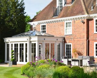 a white orangery by David Salisbury attached to a brick home, with grey rattan furniture set and manicured lawn