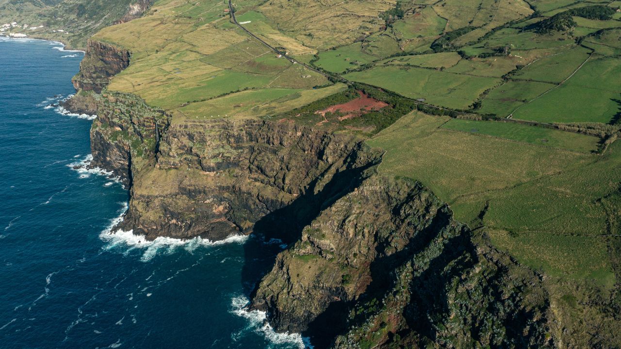 a blue ocean crashes against the bottom of a high cliff that gives way to green fields stretching to mountains in the distance.