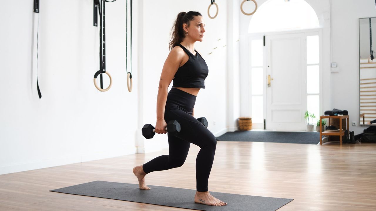 Woman doing dumbbell lunges in an open room on an exercise mat