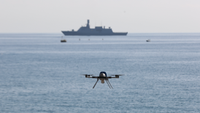 a flying naval drone with a ship in the background