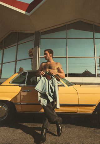 Tell Me Lies star Jackson White poses in front of yellow vintage car wearing a denim jacket, jeans, and boots.