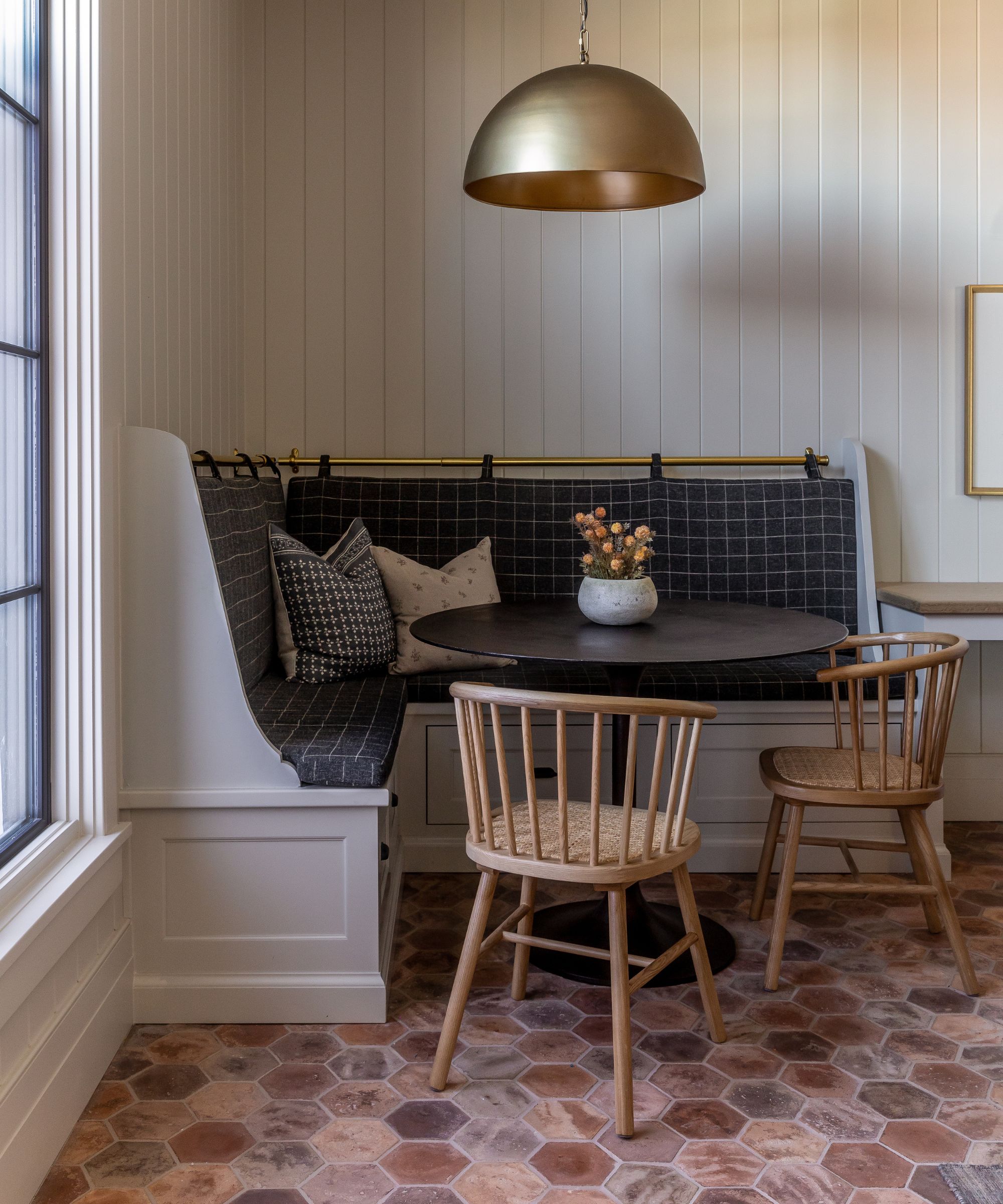 neutral panelled kitchen corner with a built in custom corner banquette seat with plaid navy cushions, a dark round table, wooden chairs and a terracotta floor tile