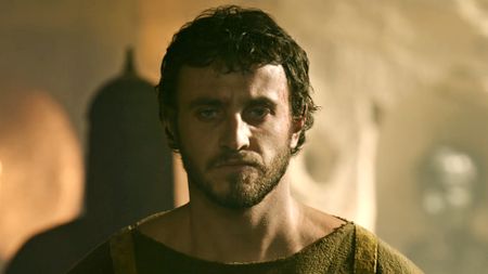 Paul Mescal, wearing a cloth tunic stands stoic, guard behind him, in Gladiator II.