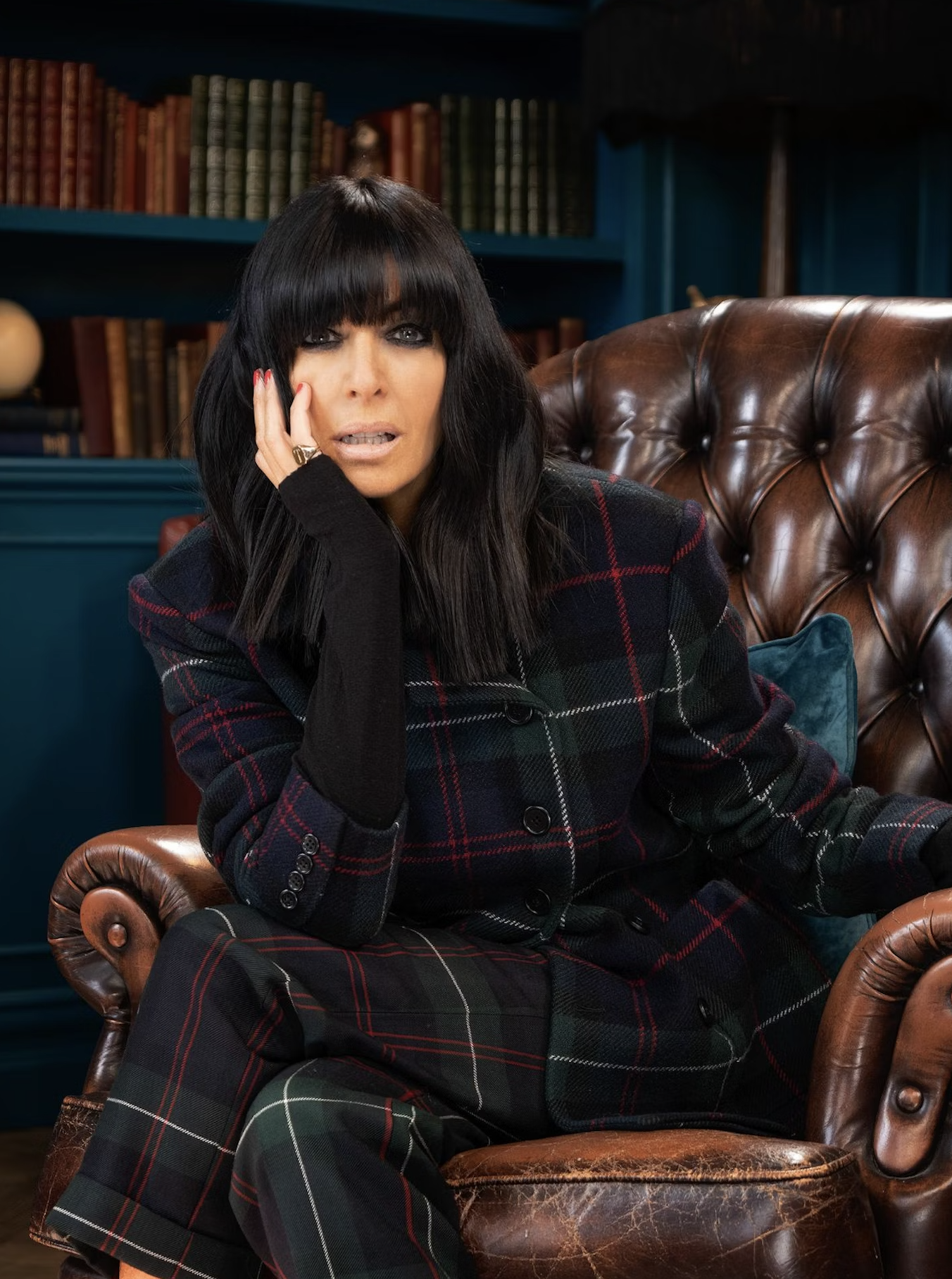 Claudia Winkleman sitting on a leather chair, wearing a checked suit