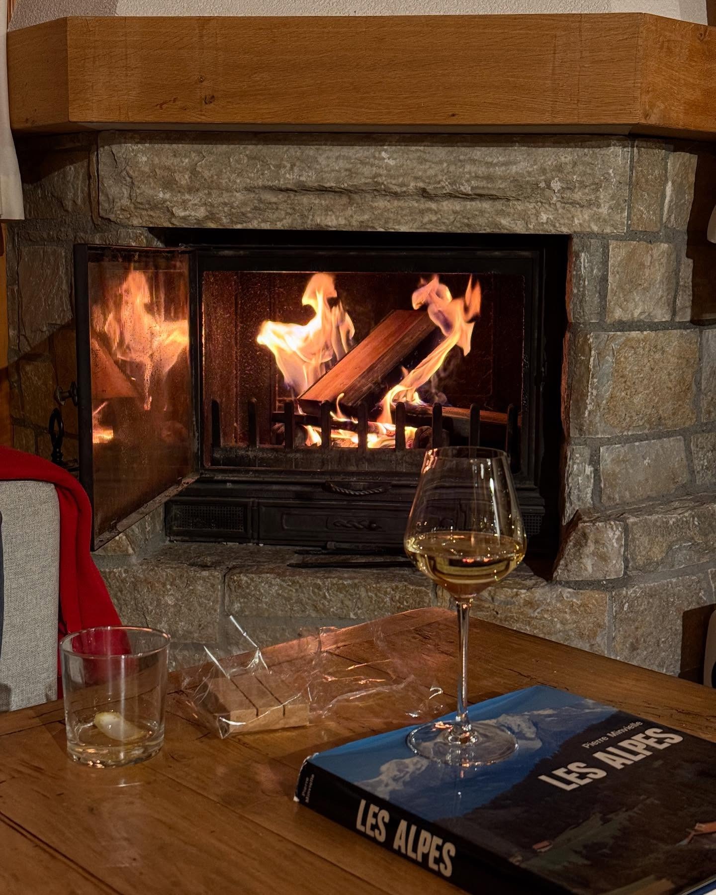 A stone fireplace scene with burning logs and a table with a glass of wine
