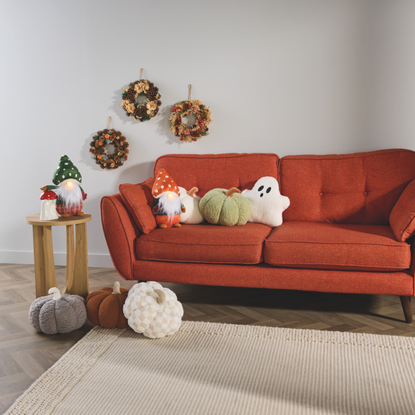 Aldi autumnal decor on a red sofa. There are plush pumpkins, ceramic lights, a ghost pillow, gons and wreathes.