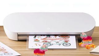 A Silhouette Portrait 4 cutting machine sits on a wooden surface, processing a sheet of paper printed with colorful wedding-themed illustrations alongside small, fringed llama pi&ntilde;atas.
