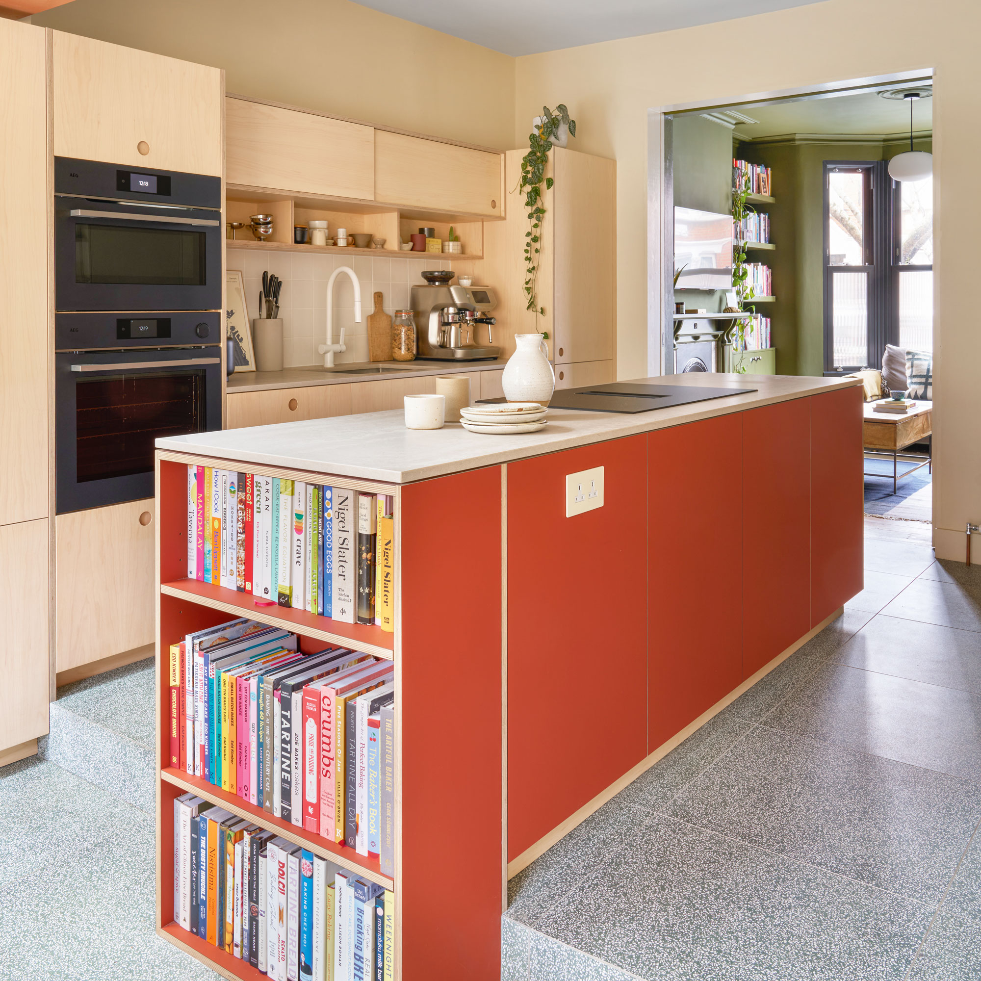 Plywood kitchen with no handles and a terracotta kitchen island with a built in bookshelf at the end of the island.