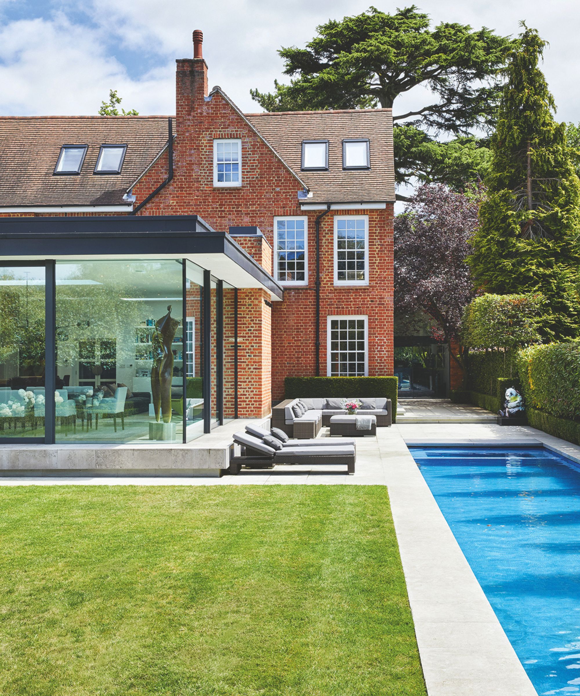 A red brick house seen from behind with a large glass conservatory, mown lawn, sun loungers, pale stone paving and a large and narrow pool