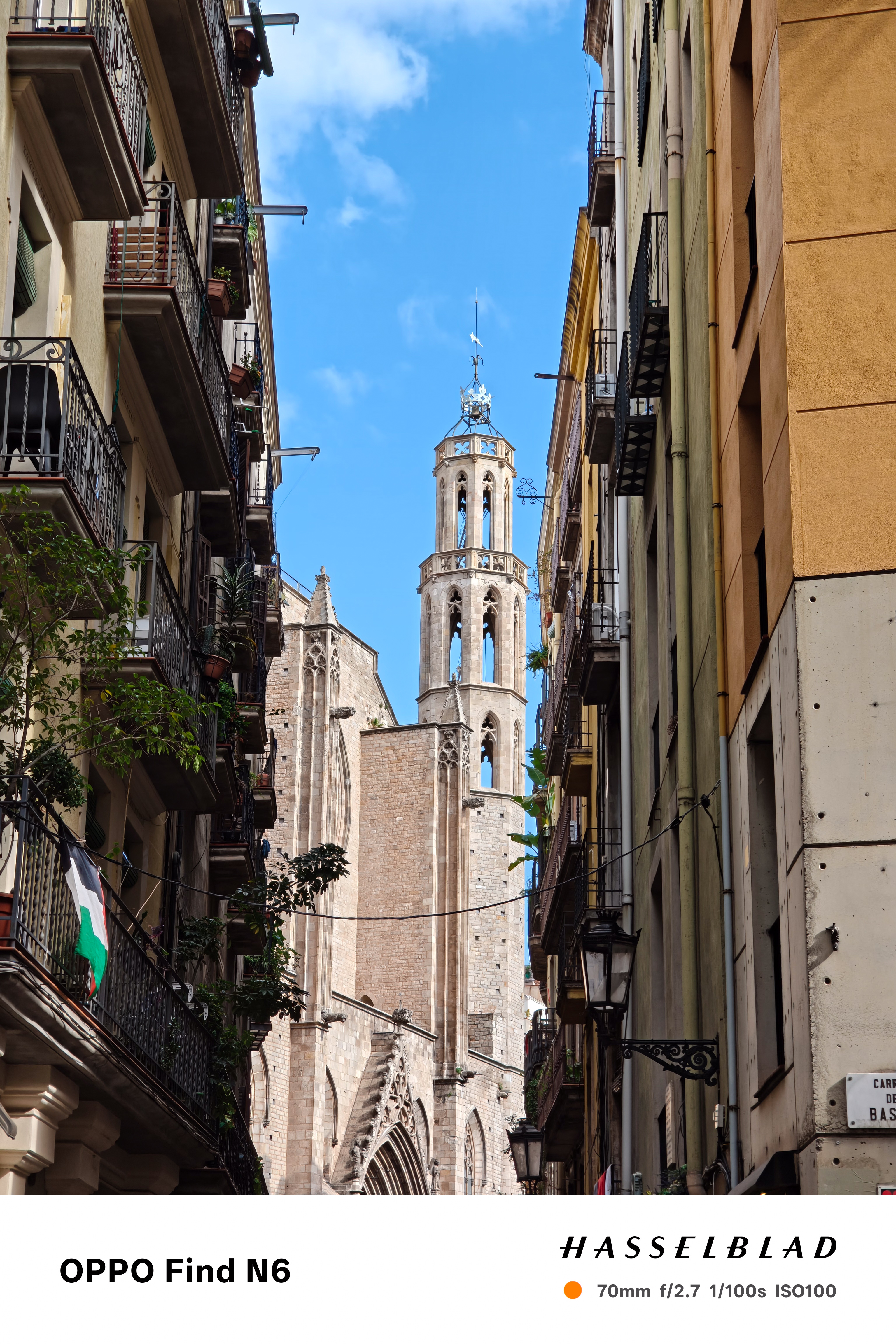 A vertical architectural shot of a narrow street in a historic European city. The view is framed by tall, rustic apartment buildings with balconies, leading the eye to a tall, intricate stone cathedral bell tower standing against a blue sky with soft white clouds.
