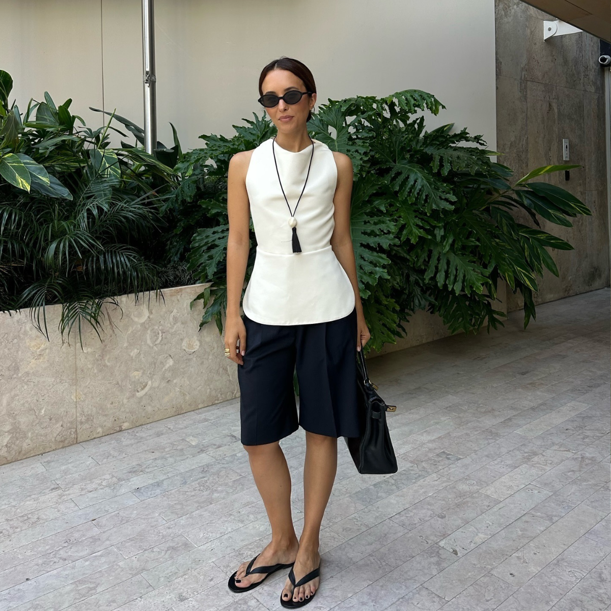 Stylist Debora wears the Reformation Jessie thong Sandal in black leather with black bermuda shorts, a white scooped neck of the shoulder top a long black frayed pendent and 90s style oval sunglasses. she is carrying a black suede bag and is stood in front of some green palm trees in an image taken from her Instagram recently.
