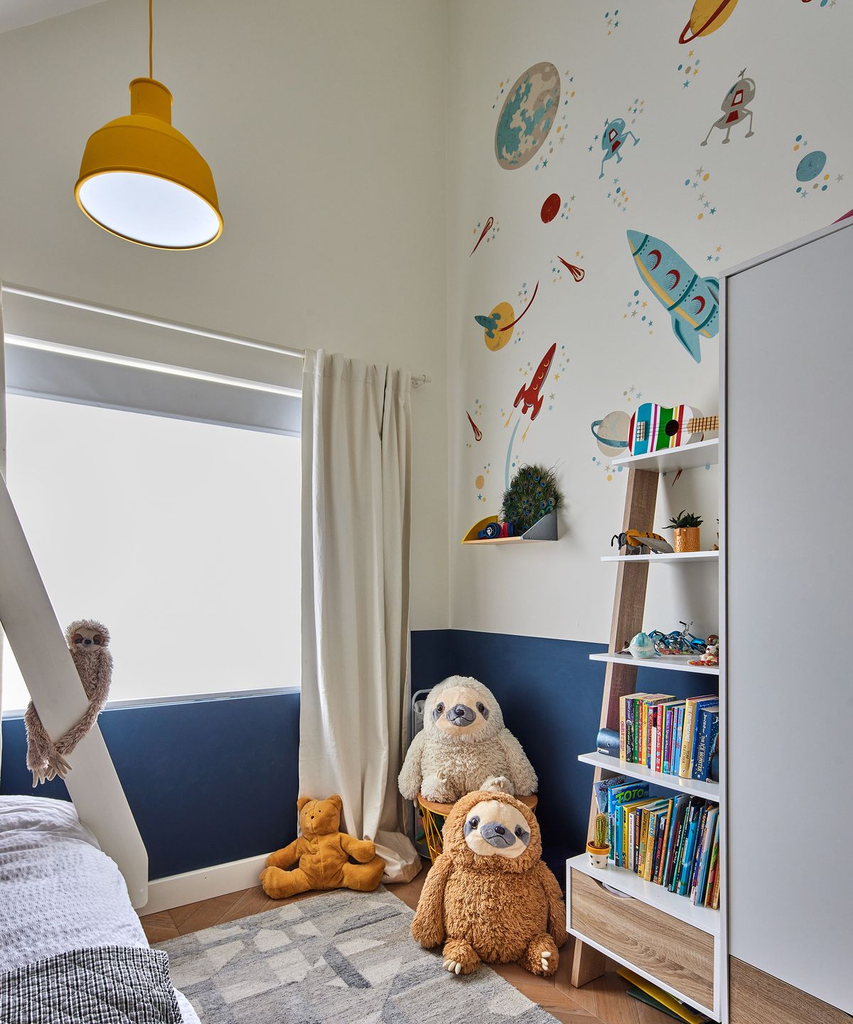 A child's room with colourful rocket wall motifs. The walls are white with a deep blue stripe. The lampshade is yellow and there are cuddly sloth toys