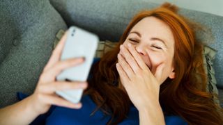 Woman laughing at video on phone, lying down on sofa at home