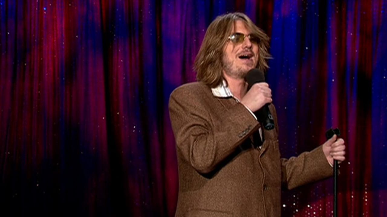 Mitch Hedberg wearing a brown sport coat and sunglasses while performing.