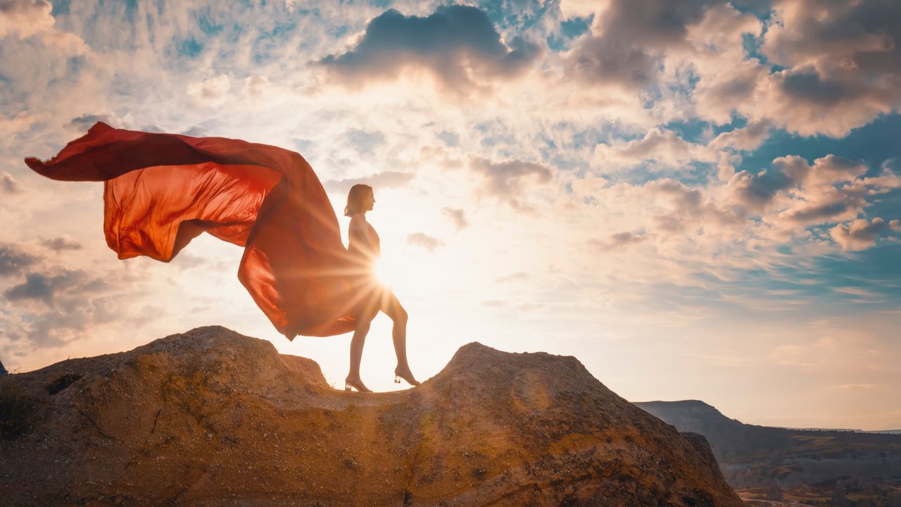 A woman in a long red dress billowing behind her like a cape, standing on a clifftop or mountaintop at sunrise 
