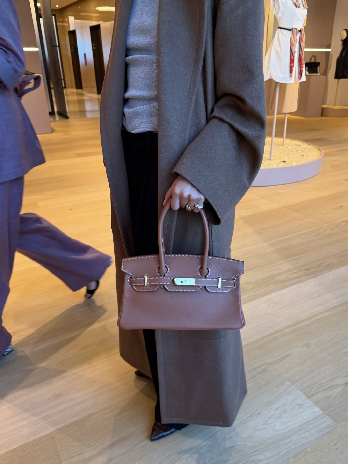 A woman wearing a mole brown coat carrying an Hermes handbag