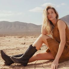 A female model sitting in the desert in a tan dress and dark brown cowboy boots.