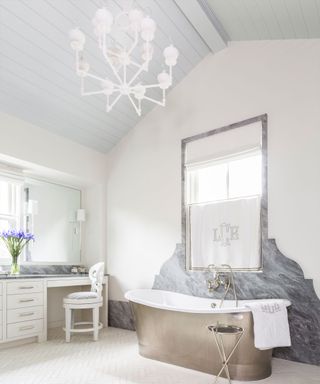 Light, bright, white bathroom with pale blue painted wooden ceiling, large framed artwork and marble design on wall beside metal bath, white chandelier, vanity area with mirror, storage, vase of flowers, chair, white tiled floor