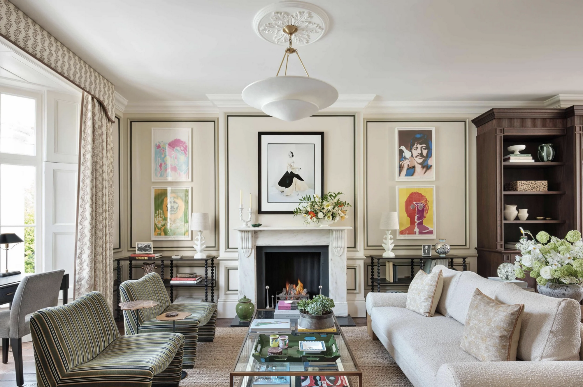 living room with ornate fireplace with wall moulding with artwork, pendant light, over white sofa with green accent chairs opposite a glass coffee table, with patterned curtains with a matching pelmet