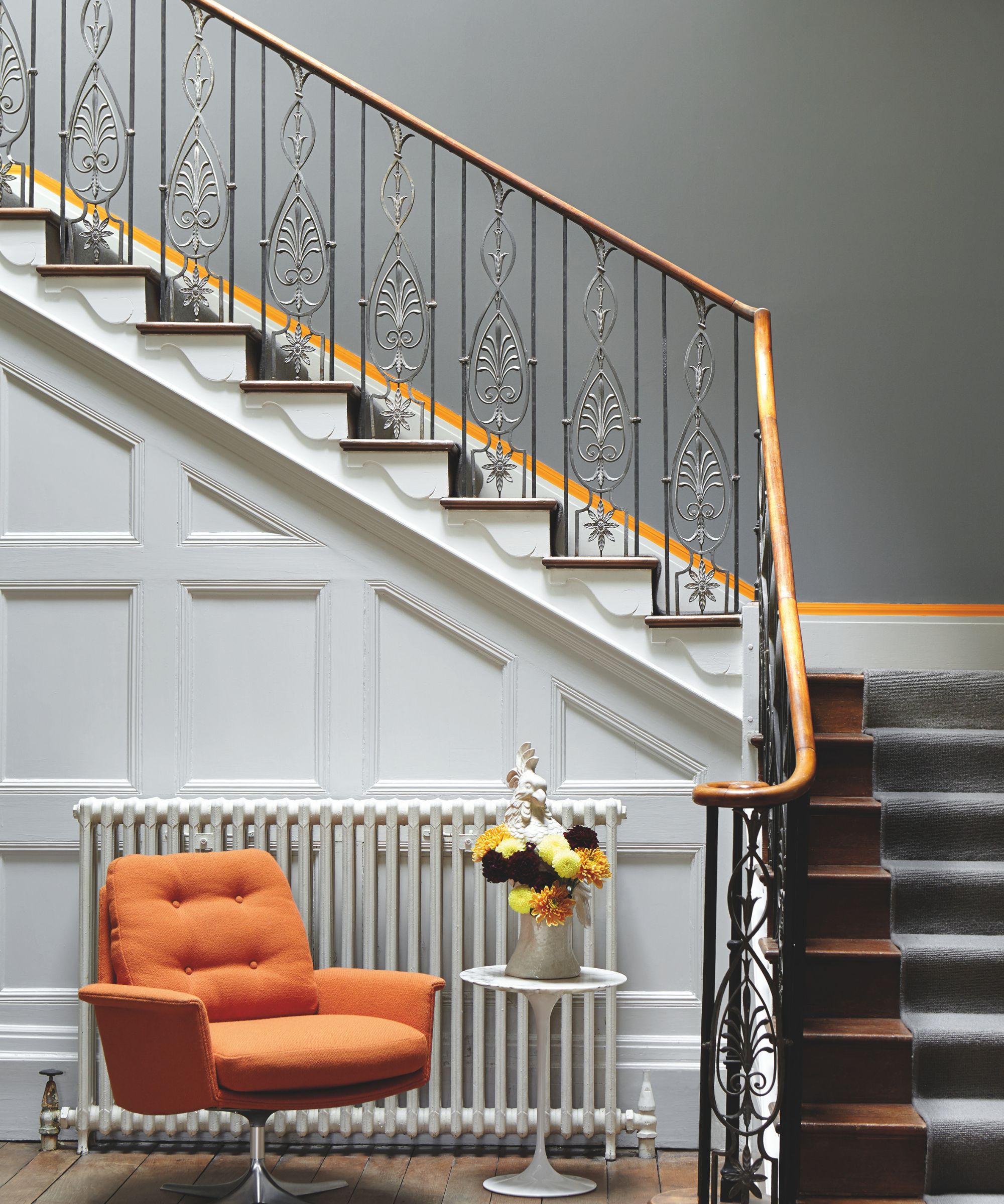 Staircase with grey walls, iron railings, an orange painted line on the wall, white panelling below with a white radiator in front of it and an orange armchair beside a white round side table