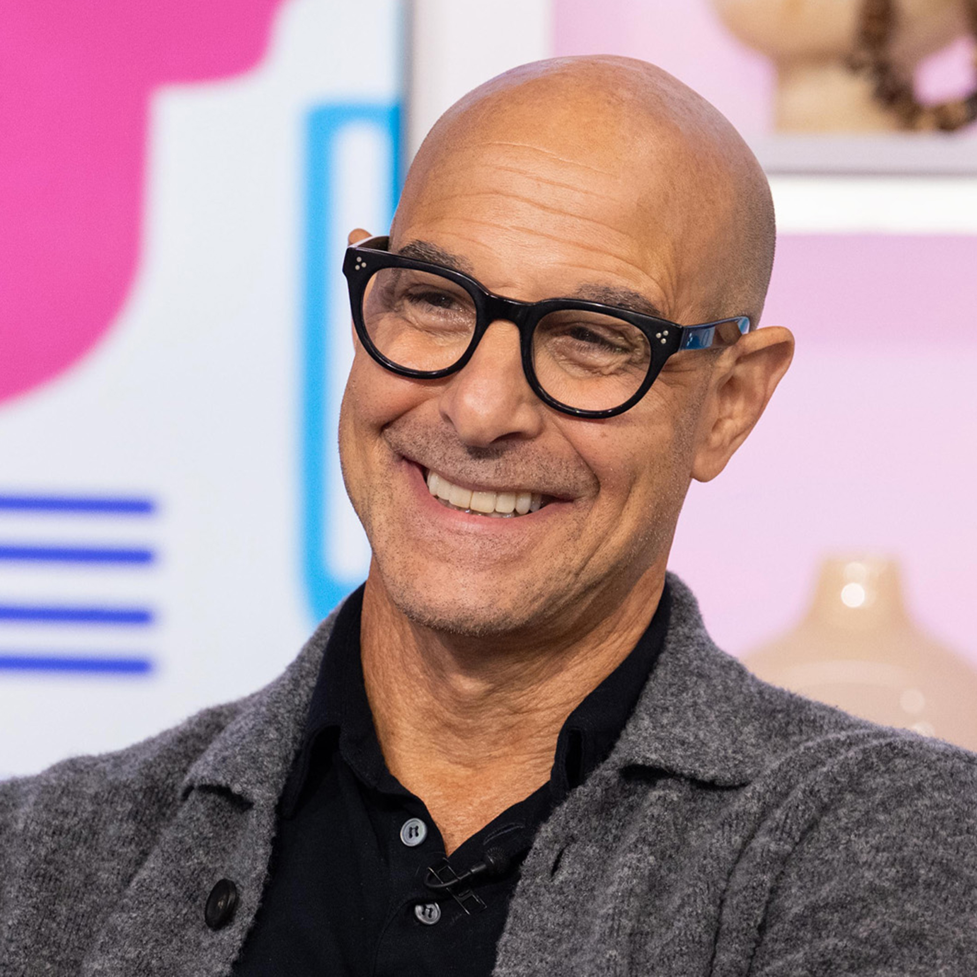 Stanley Tucci wearing gray and black in front of pink and blue cooking background