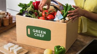 A hand reaches into a Green Chef meal kit filled with fresh vegetables