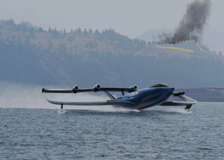 View of Marcus Lawson's blue concept fire-fighting plane on the water with hills and black smoke in the background