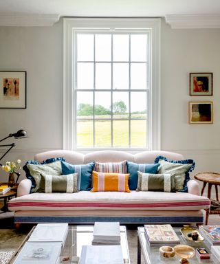 living room with neutral walls, period features, artwork, bold stripe couch with bold couch pillows, two glass coffee tables with books and objects, retro black floor lamp, rug, view outside